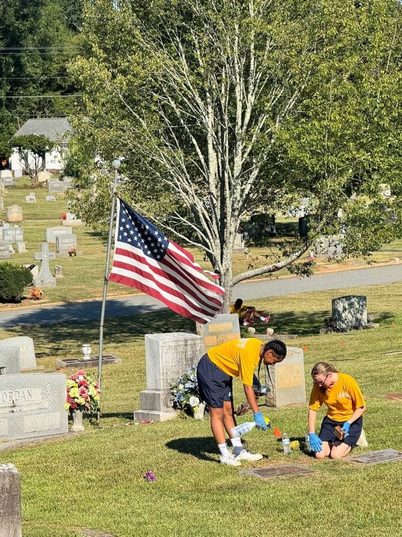 Two Naval JROTC cadets kneel to clean a veteran's grave marker by hand at Greenwood Cemetery.